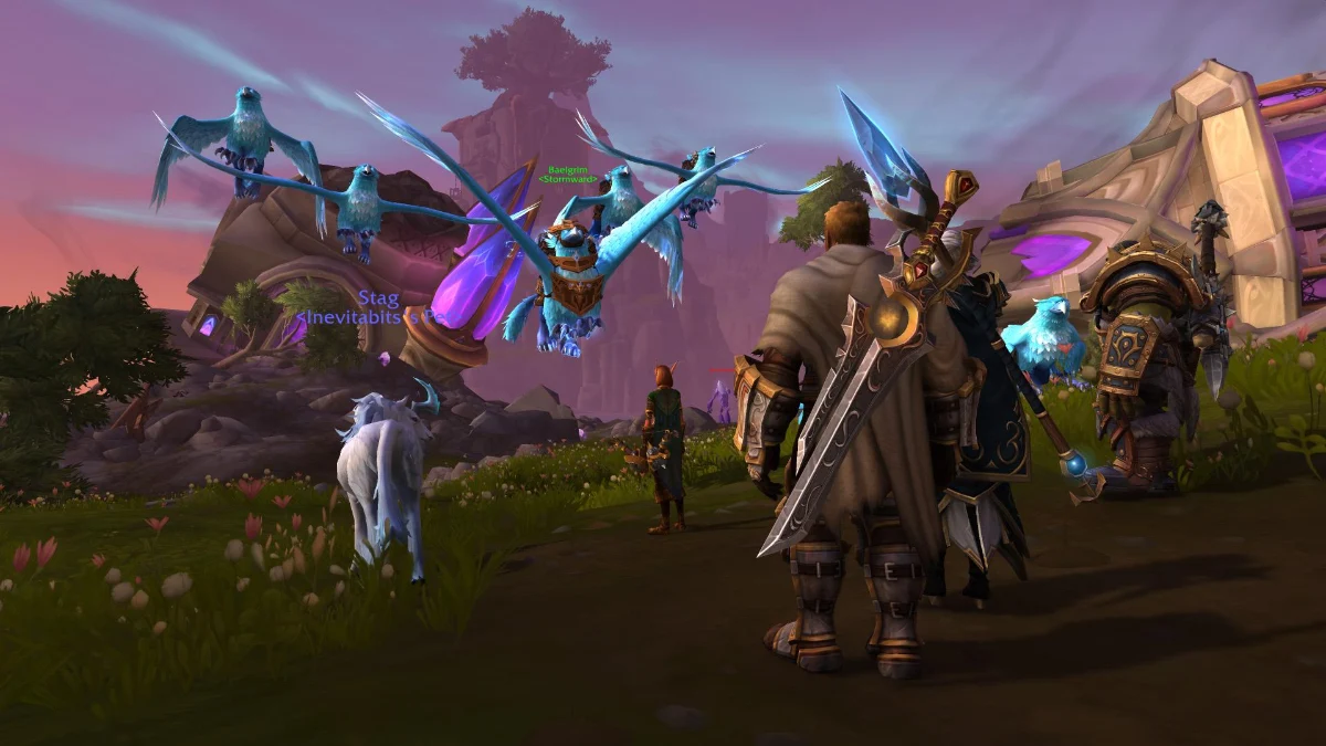 Three players in fantasy armor stand in a grassy field with pink flowers, facing a magical structure with purple crystals. Two blue, winged bird-like creatures fly in the background, and a white, horned creature grazes nearby.