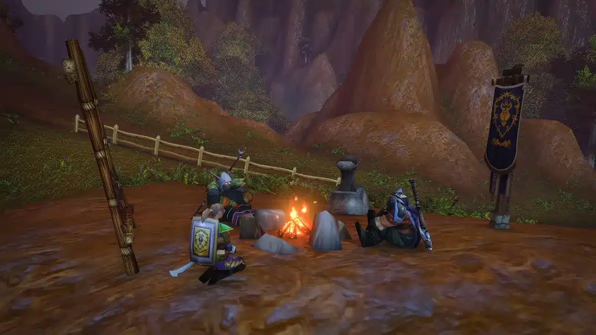 A trio of characters sit around a campfire in a wooded, hilly landscape, with a banner bearing a crest visible in the background.